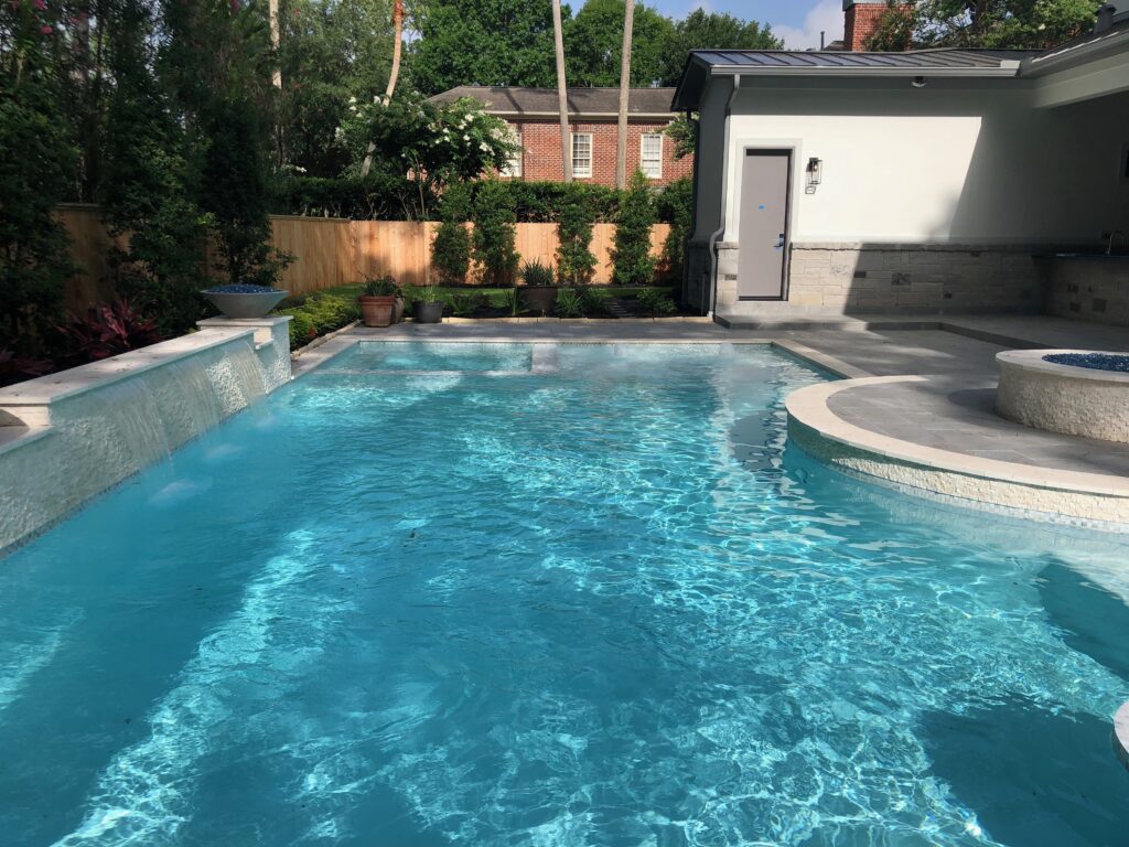 Luxury Custom Gunite Pool Design: Raised Beam Feature Wall, Flush Spa, and In-Pool Dining Oasis Luxury custom in-ground gunite pool in Memorial Villages Texas with a stacked stone raised beam wall, integrated in-pool dining table, flush-mount spa, and a poolside fire pit.