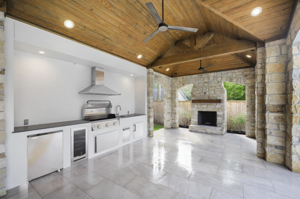 Large luxury patio cover in Houston featuring a stained vaulted tongue and groove ceiling, stone facade outdoor kitchen, fireplace, and honed grey tile flooring.