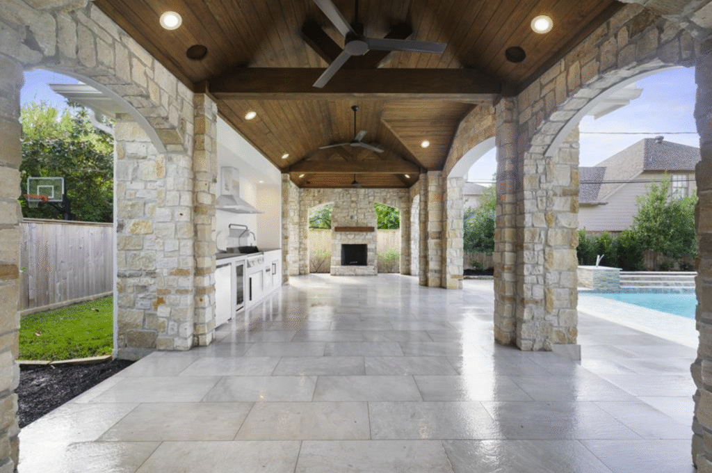 A custom patio cover in Tanglewood featuring a stained tongue and groove ceiling, stone facade, and an outdoor kitchen with a fireplace, featured in our luxury outdoor living portfolio in the Houston area.