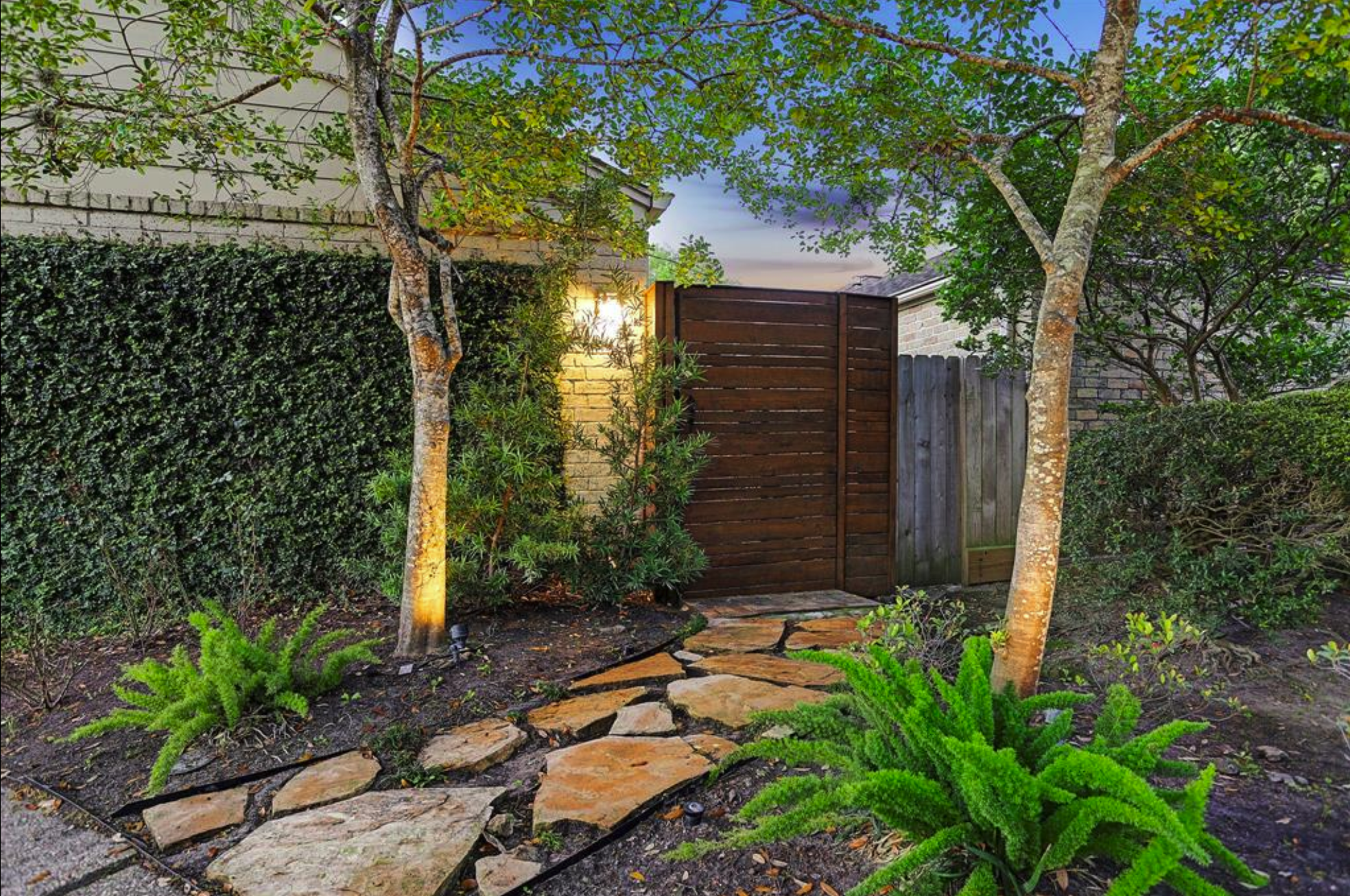 A modern horizontal stained wood fence gate with a natural flagstone path surrounded by lush landscaping and lighting.