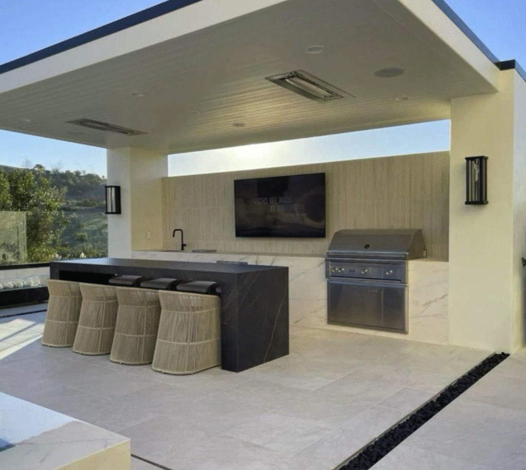 A luxury outdoor kitchen featuring a built-in grill, sink, and island bar seating underneath a premium modern 4K Aluminum patio cover with an integrated privacy wall, showcasing elite outdoor kitchen and fireplace designs.