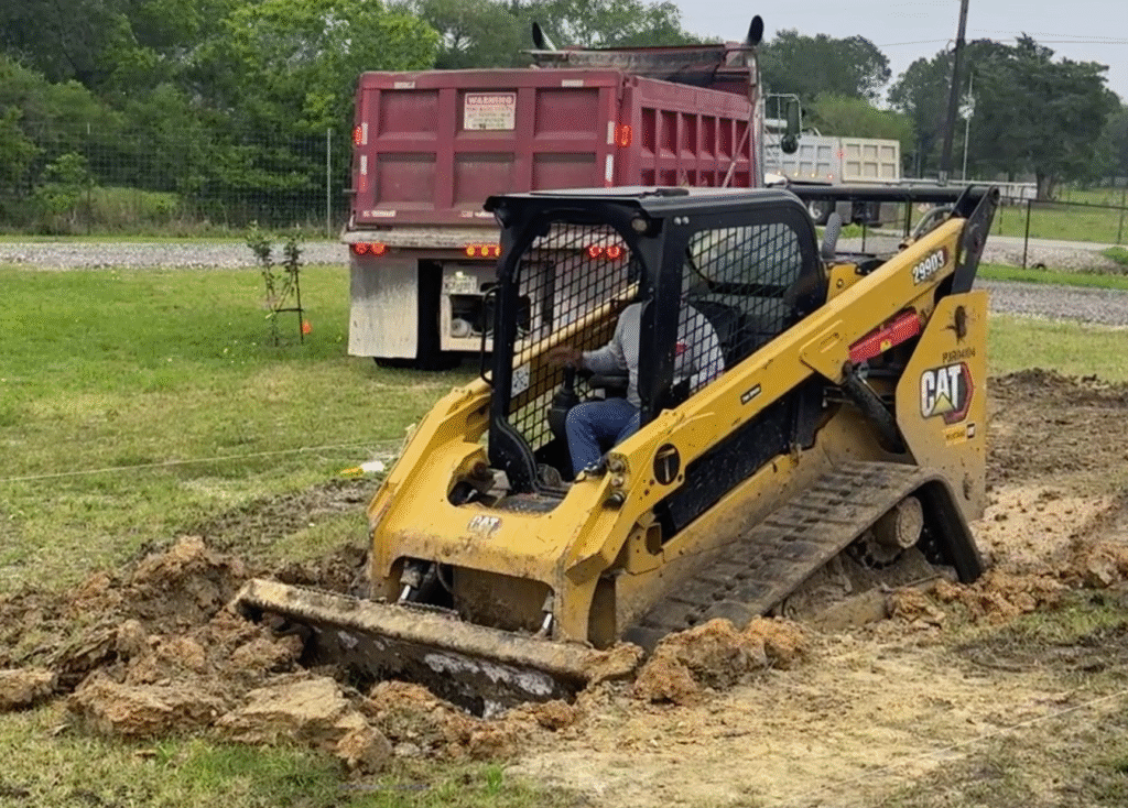 In-Ground Swimming Pool Excavation: Heavy Equipment Digging Custom Backyard Pool Shell Residential swimming pool excavation service in Houston Texas area featuring a yellow hydraulic excavator digging a dirt pit with tandem dump trucks hauling soil.