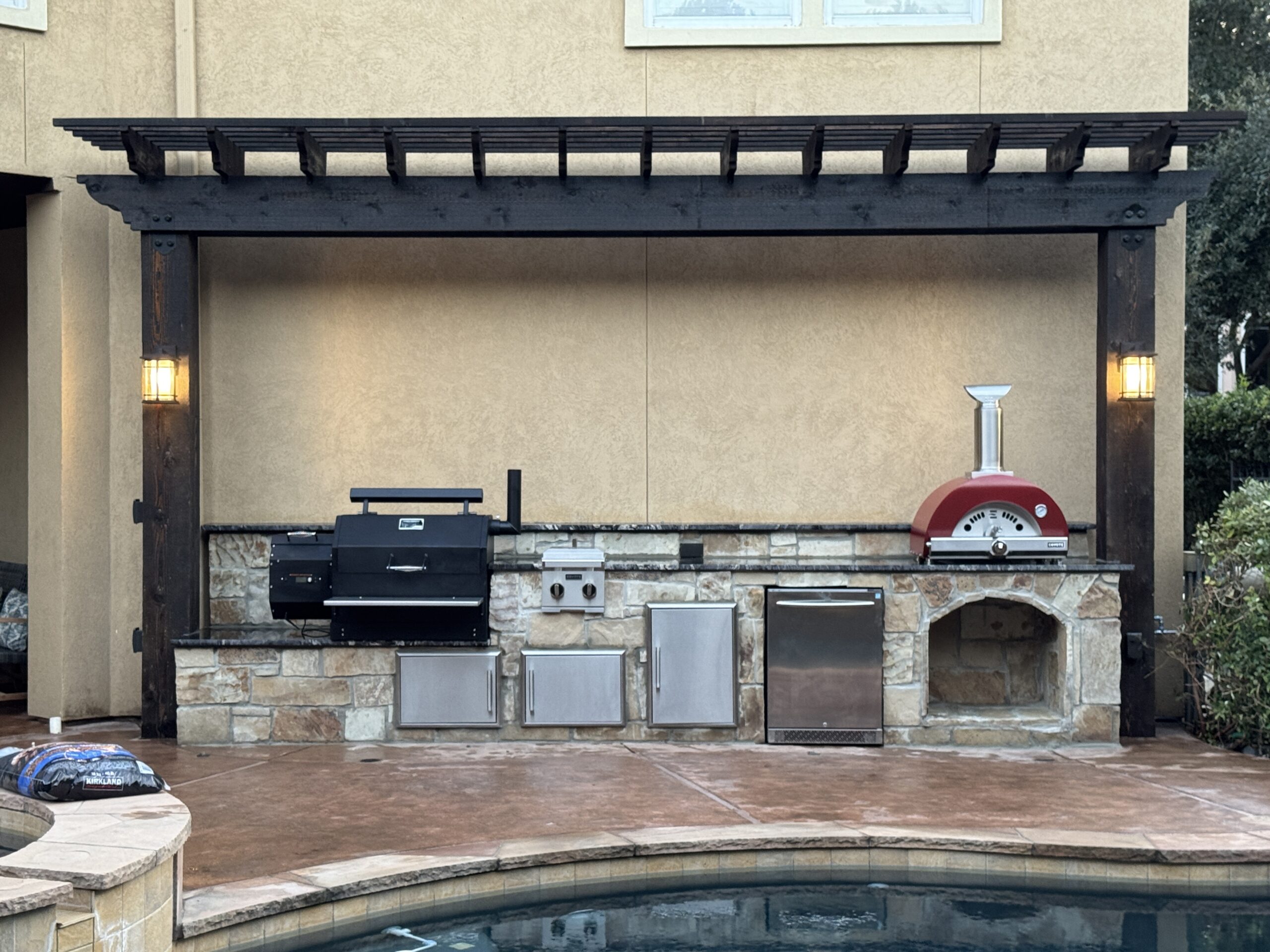 Located in Sugar land this outdoor kitchen has a stone facade under a dark-stained pergola, featuring a Yoder pellet grill, Coyote pizza oven, gas burner, built-in refrigerator, trash drawer, and integrated wood storage.