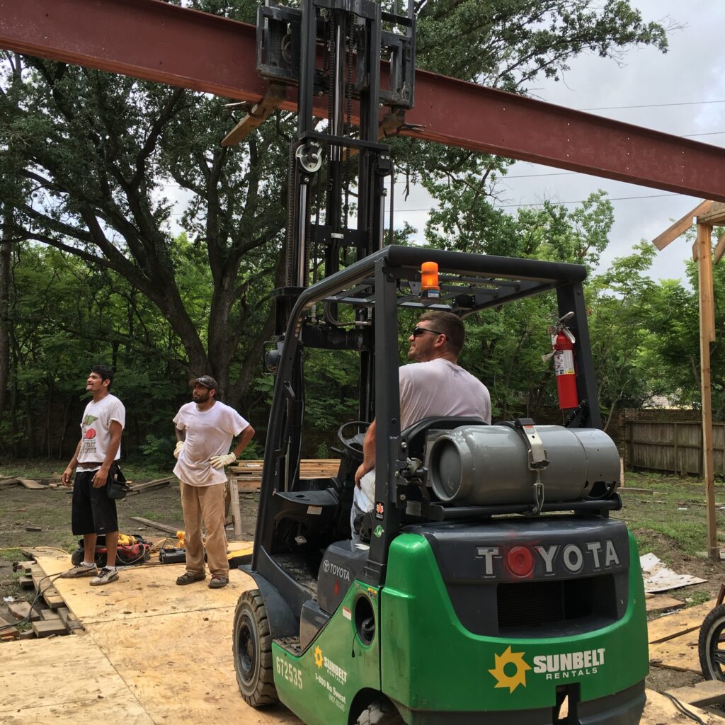 Expert Outdoor Building & Implementation | Houston Outdoor Living Builders A framing crew maneuvers a heavy structural steel I-beam into place using a forklift for a large custom patio cover, demonstrating design and engineering used by premier Houston Outdoor Living Builders in Tanglewood