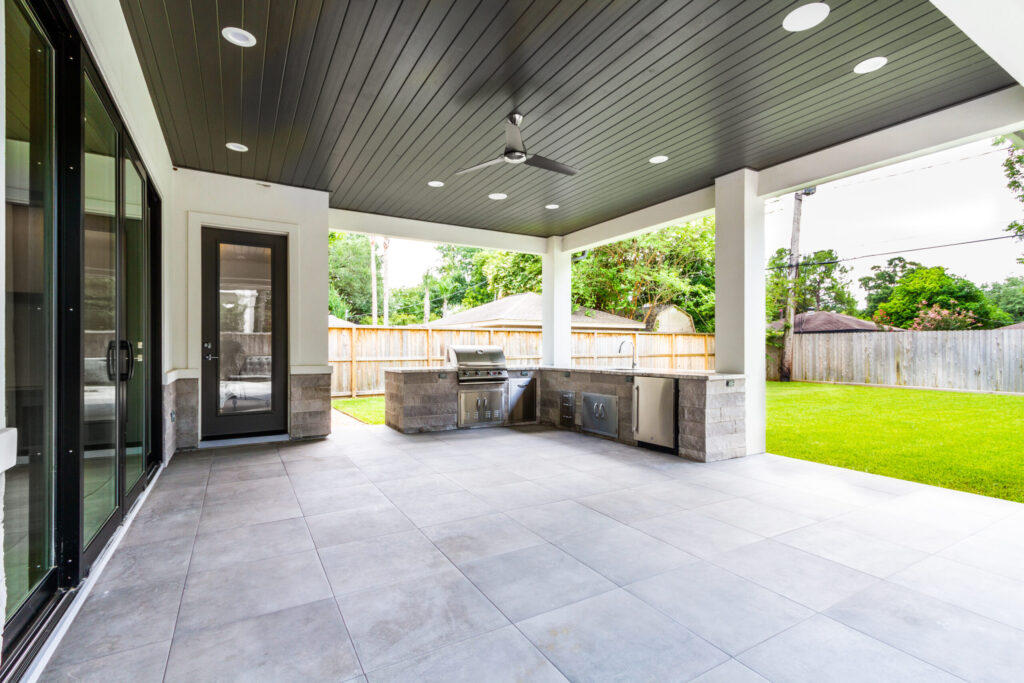 Large custom patio cover in Katy featuring structural stucco columns, a painted tongue and groove ceiling, an L-shaped outdoor kitchen with a stone facade, and tile flooring.