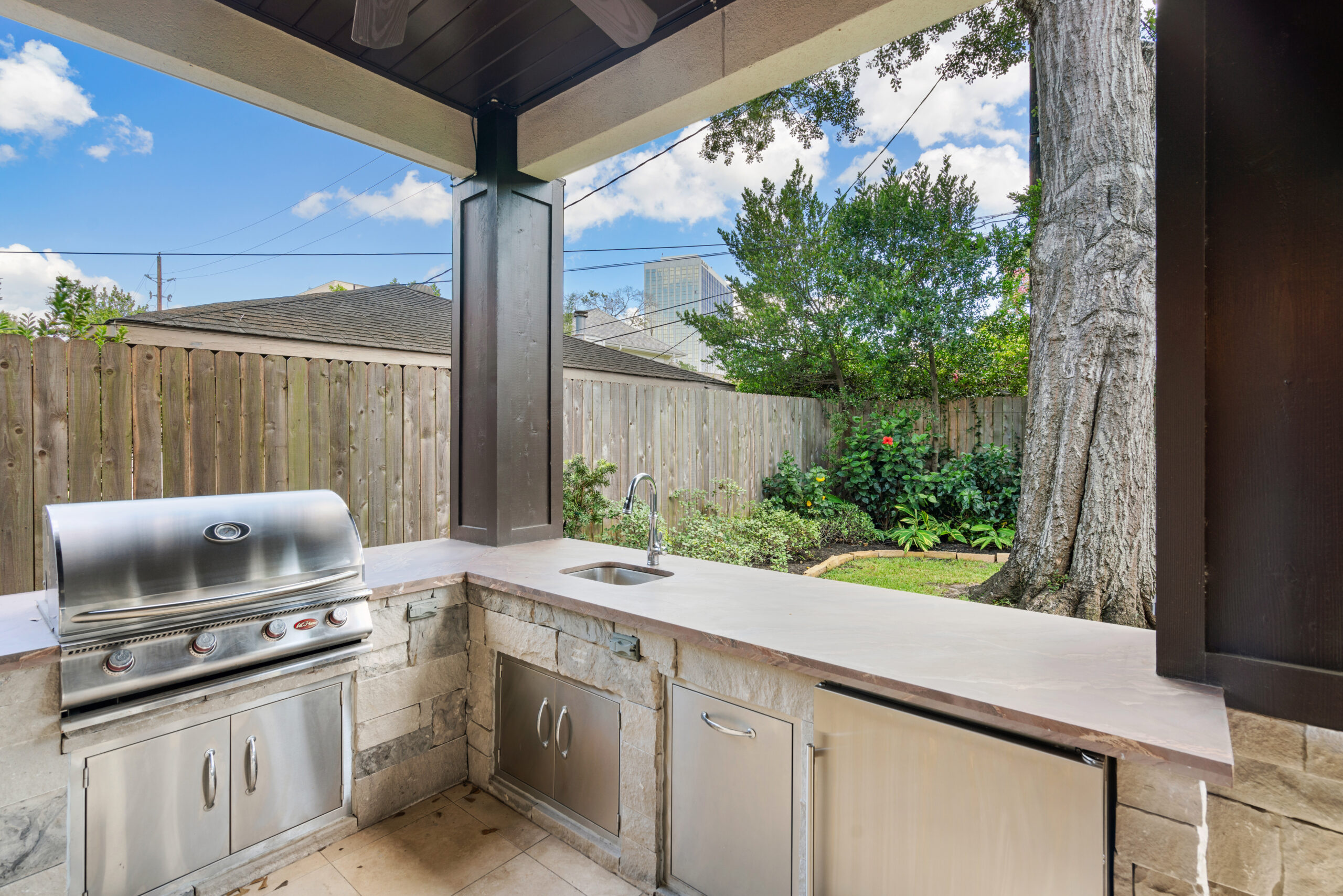 Custom stone-facade outdoor kitchen with a built-in stainless steel grill, sink, and under-counter refrigerator beneath a covered patio, built by Luxscape Outdoor Builders