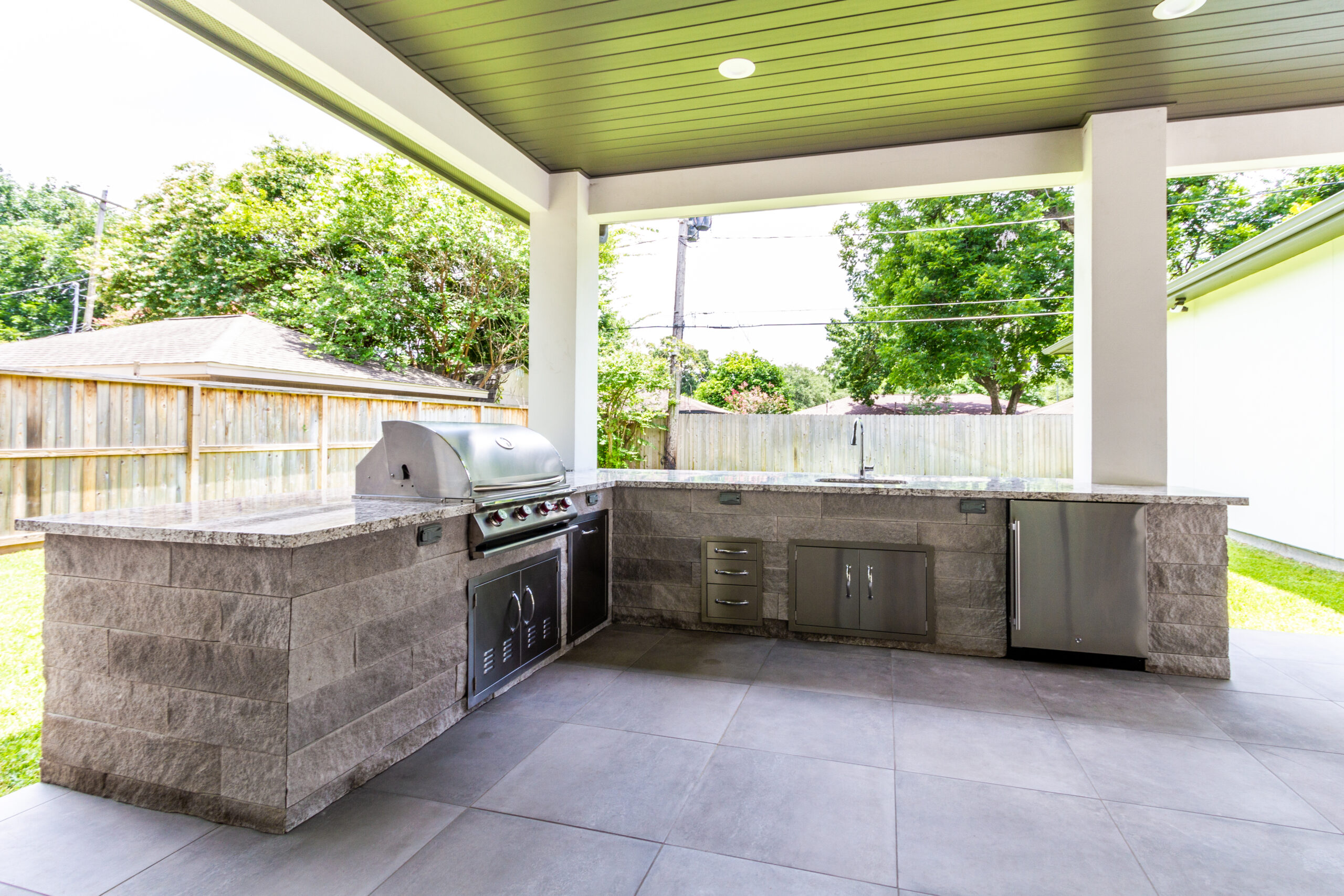 -shaped outdoor kitchen island with granite countertops, stone veneer facade, built-in stainless steel grill, side burner, and under-counter refrigerator beneath a covered patio, built by Luxscape Outdoor Builders