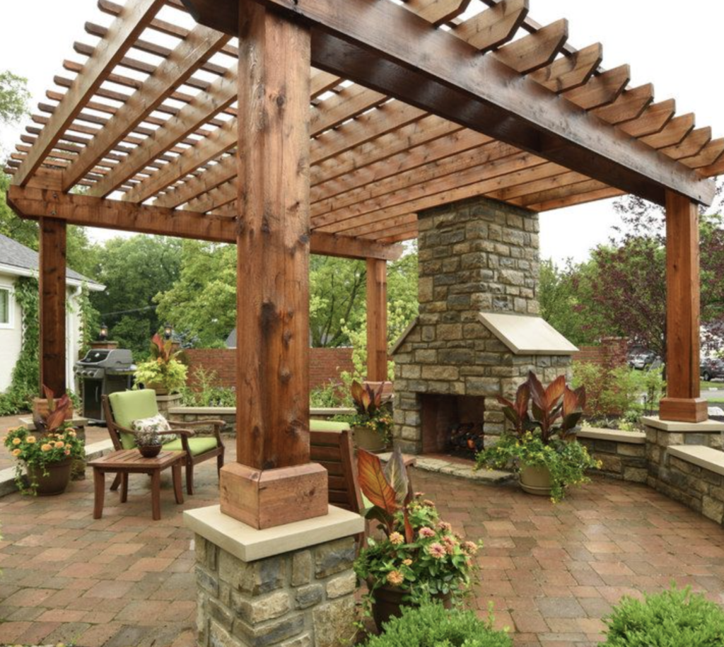 A stained cedar pergola on stone column bases beside a stacked stone fireplace, over a brick paver patio with landscaping, Luxscape Outdoor Builders serving Houston, TX.