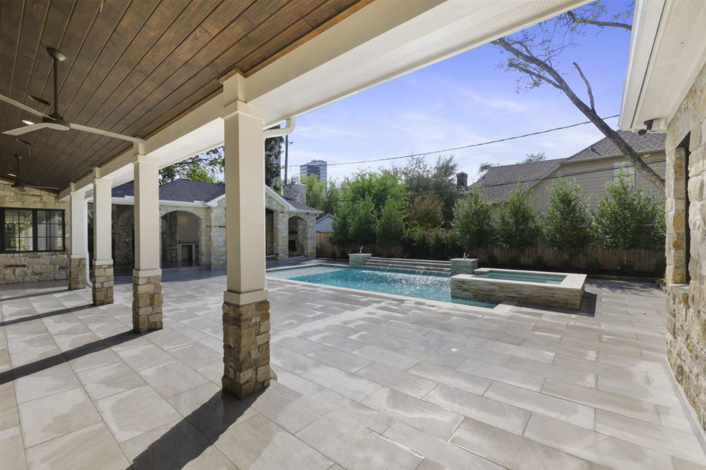 A view from a covered patio with stone column bases across a tile overlay deck to a custom pool and spa, built by Luxscape Outdoor Builders serving Houston, TX.