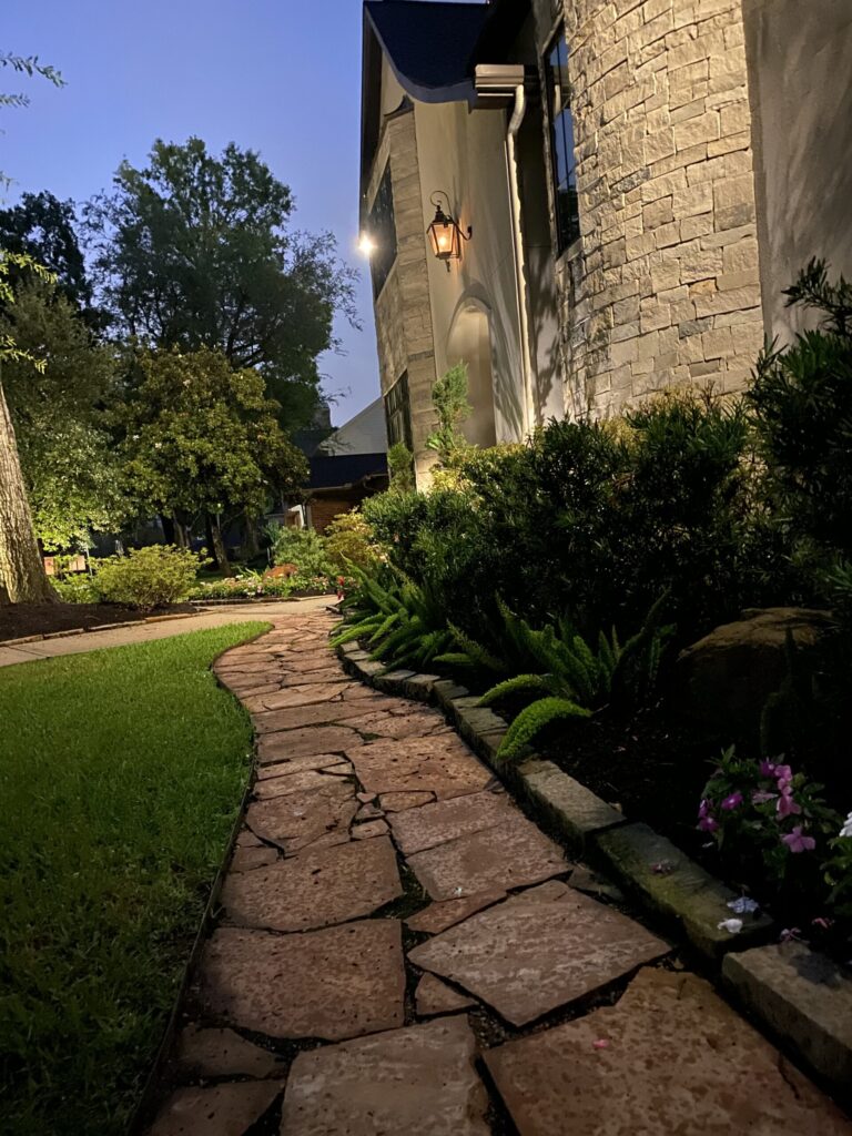 A curving flagstone pathway with stone edging, layered plantings and landscape lighting beside a stone home, built by Luxscape Outdoor Builders serving Houston, TX.