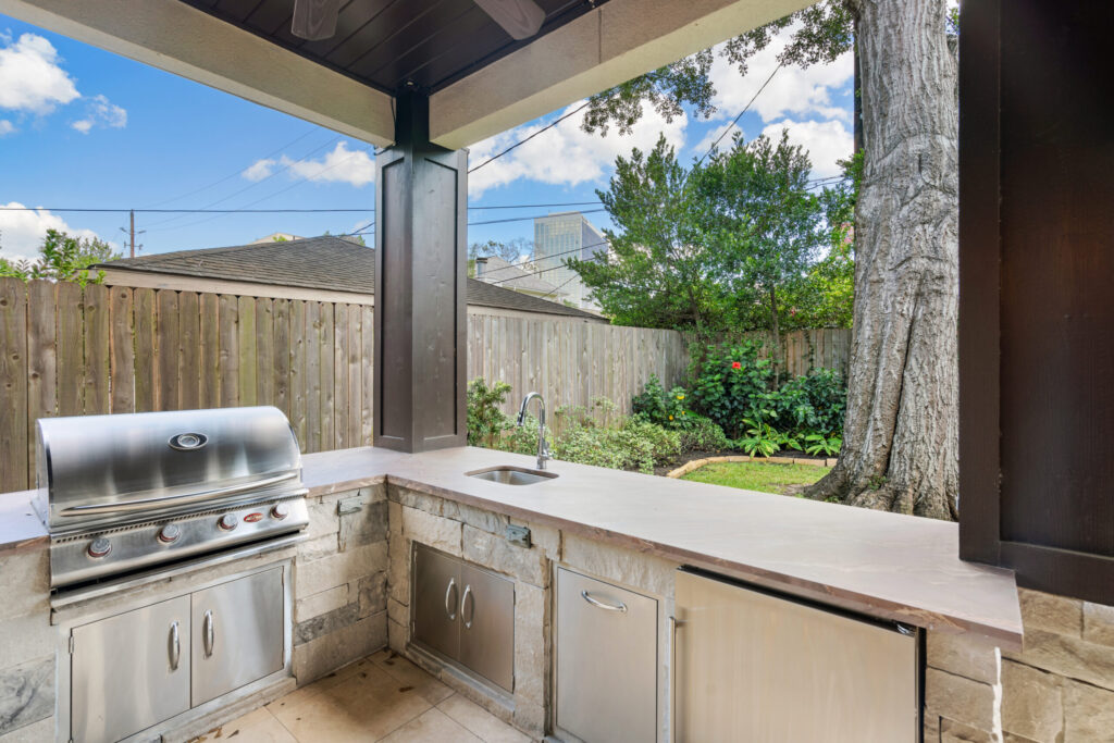 An L-shaped outdoor kitchen with a stainless steel grill, sink and storage under a covered patio overlooking a lawn and mature oak, built by Luxscape Outdoor Builders serving Houston, TX.