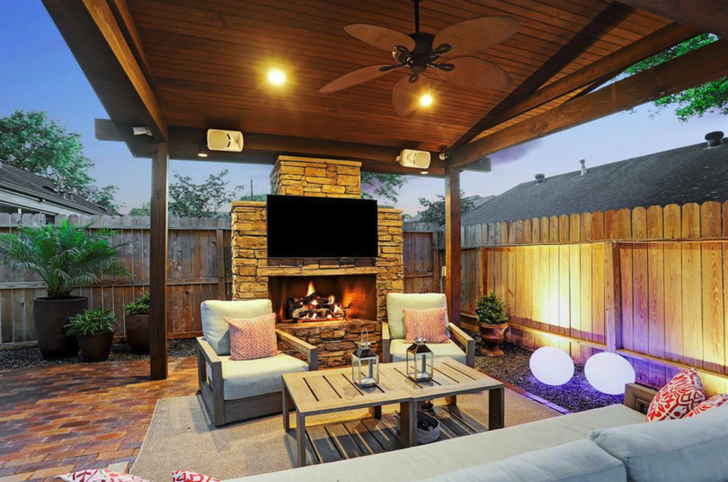 A stacked stone outdoor fireplace with a mounted TV under a stained cedar patio cover with a ceiling fan and lounge seating, built by Luxscape Outdoor Builders serving Houston, TX.